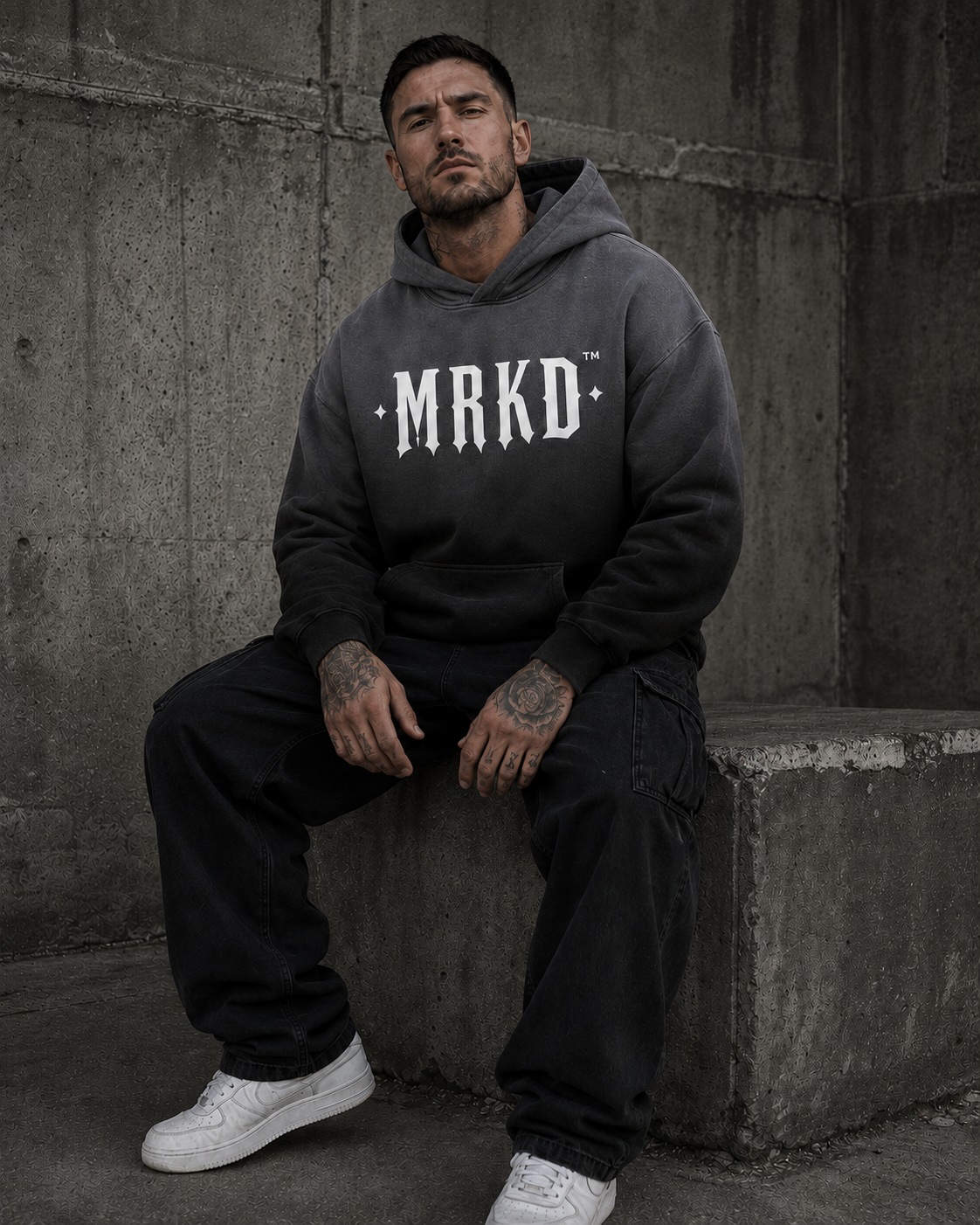 A man wearing a dark gray oversized hoodie with 'MRKD' printed in white across the chest, paired with black cargo pants and white sneakers, sitting against a concrete wall.