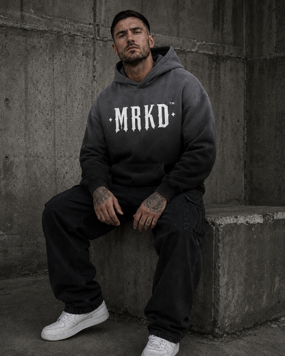 A man wearing a dark gray oversized hoodie with 'MRKD' printed in white across the chest, paired with black cargo pants and white sneakers, sitting against a concrete wall.